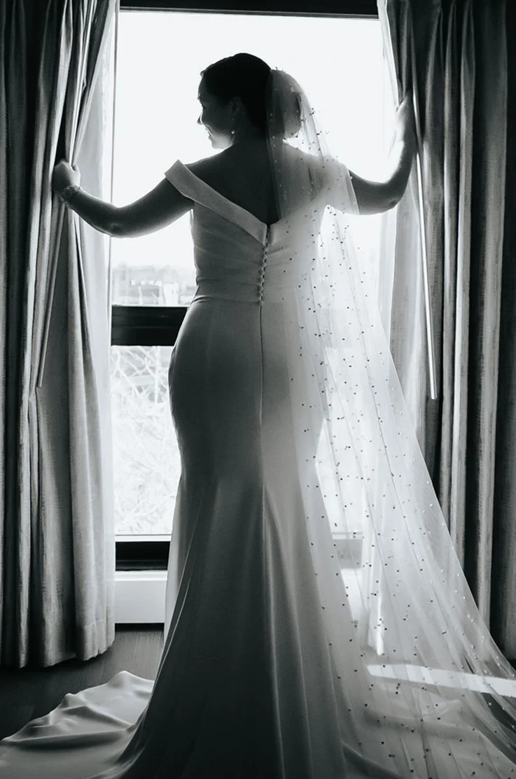 Back of bride at a window with open curtains, in a V-shaped back crepe gown with a fit and flare skirt and a pearl edge veil.