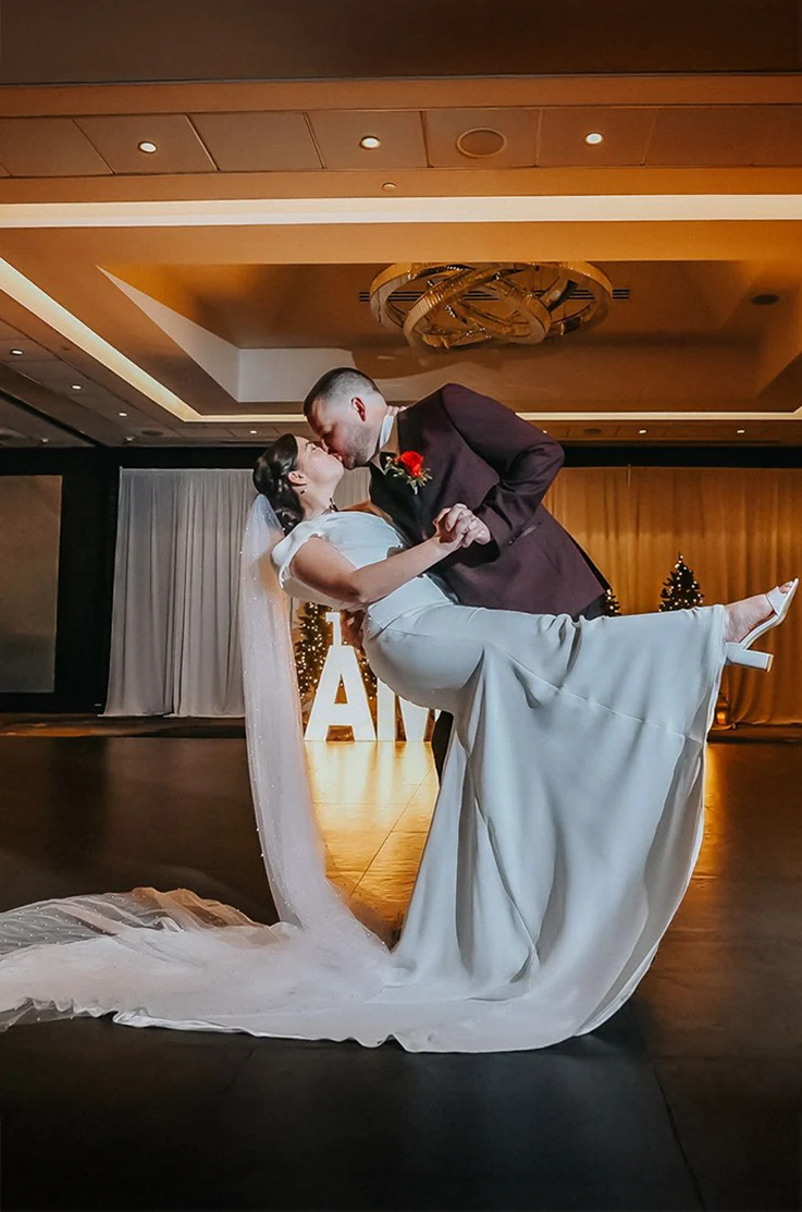 Groom dips his bride in a crepe fit and flare wedding dress with a long pearl veil and they kiss in a grand ballroom.