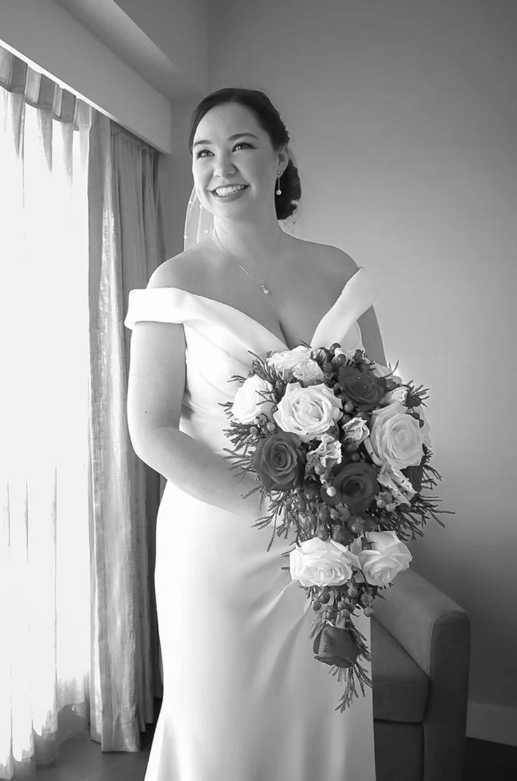 Smiling bride in a crepe V-neck fitted gown, holding a large bouquet in a bridal suite by a bright window, in black and white.