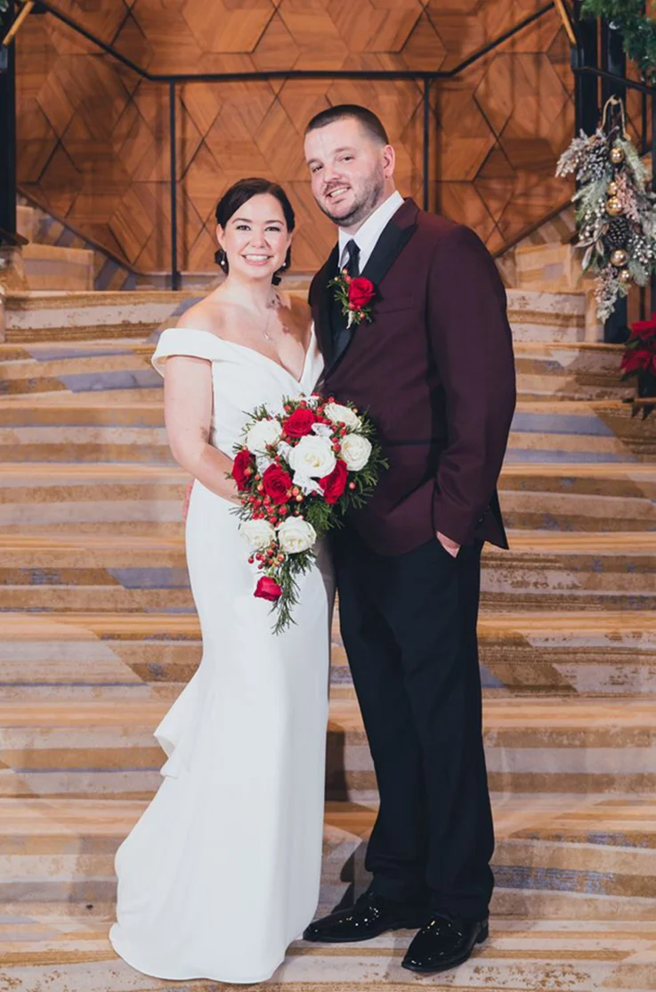 Bride in a V-neck fit and flare gown, poses alongside her groom on a grand staircase holding a white and red bouquet.