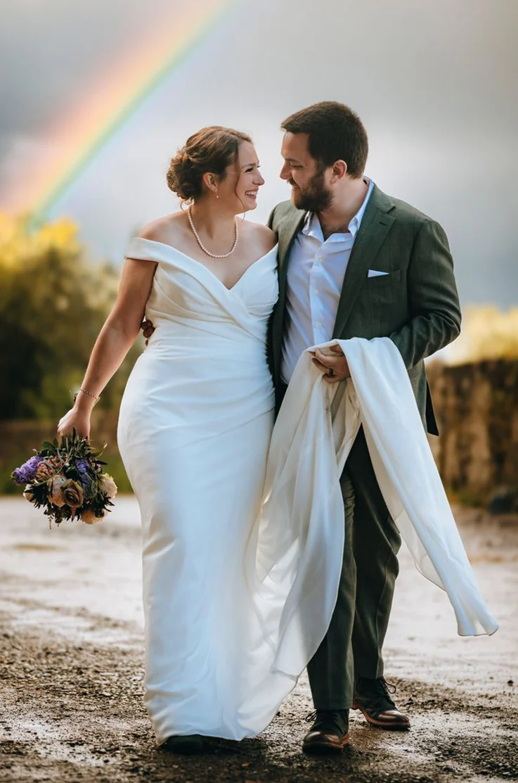 Groom holds his bride's train in a off-shoulder V-neck gown with a crepe draped bodice, smiling outdoors by a rainbow.