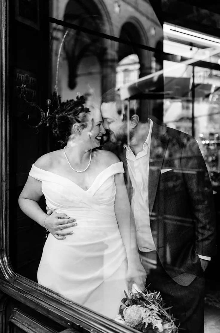 Groom embraces his bride in an off-the-shoulder crepe fitted wedding dress with draping and a V-neckline, at a window.