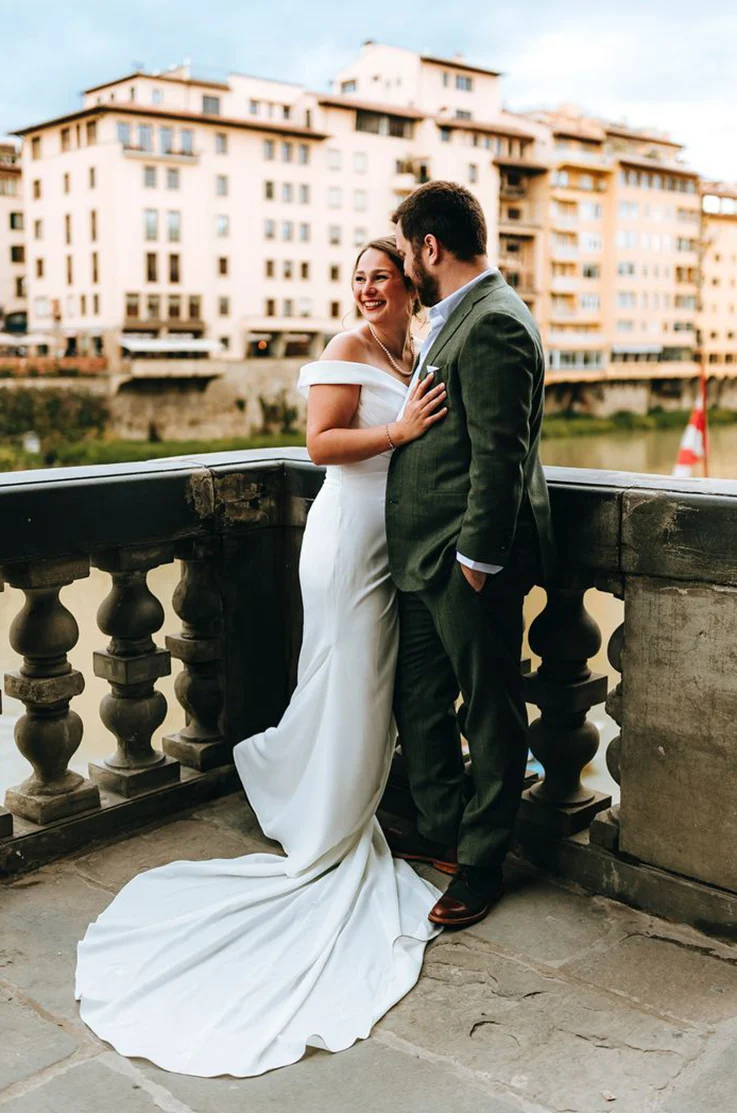 Bride in an off-shoulder crepe fitted gown with draping, and poses with her groom on a stone terrace.