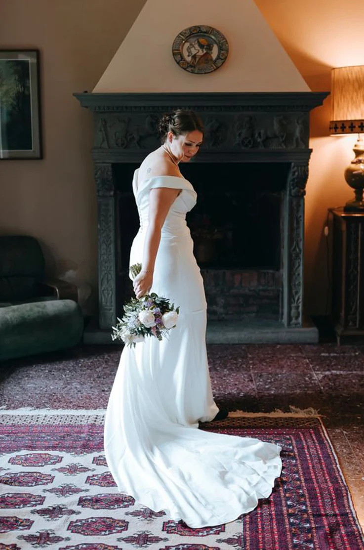 Bride looks over her shoulder in a V-shaped back crepe gown with draping and fit and flare skirt, in front of a fireplace.
