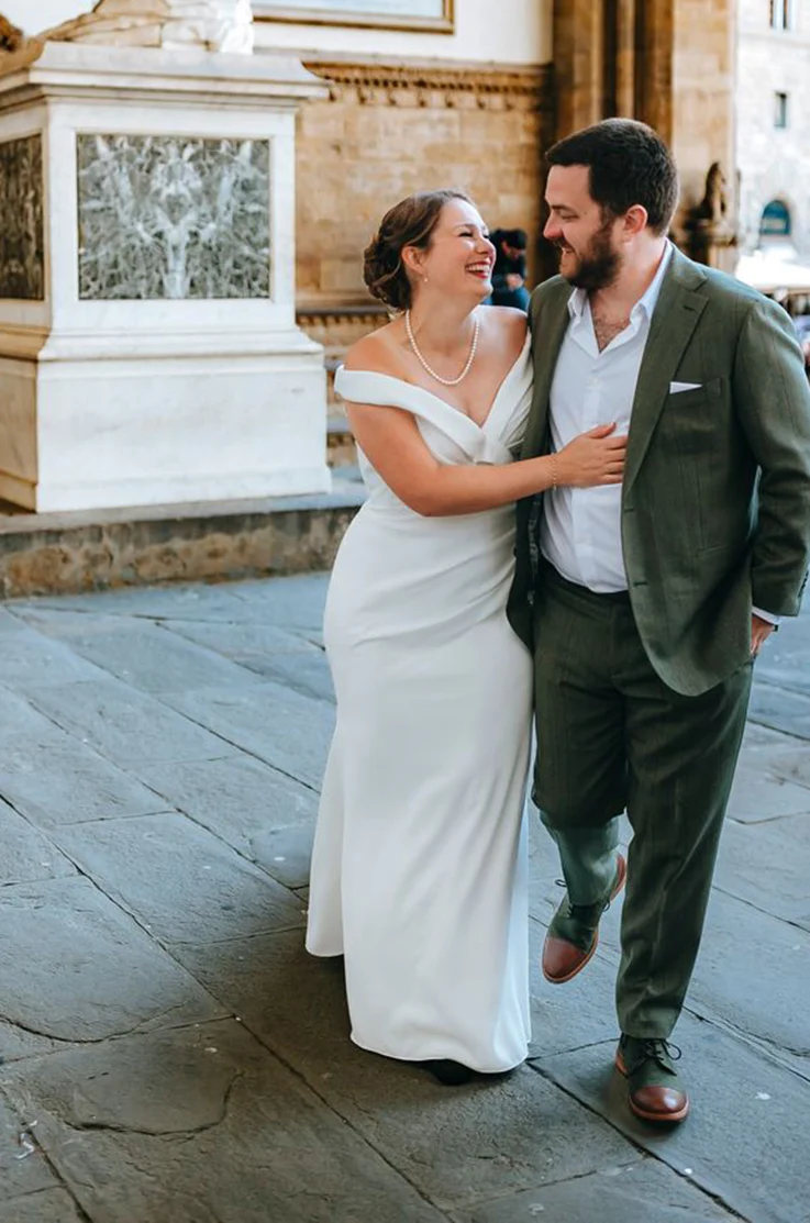 Bride in an off-shoulder fit and flare gown with a draped crepe bodice, laughing alongside her groom as they walk.