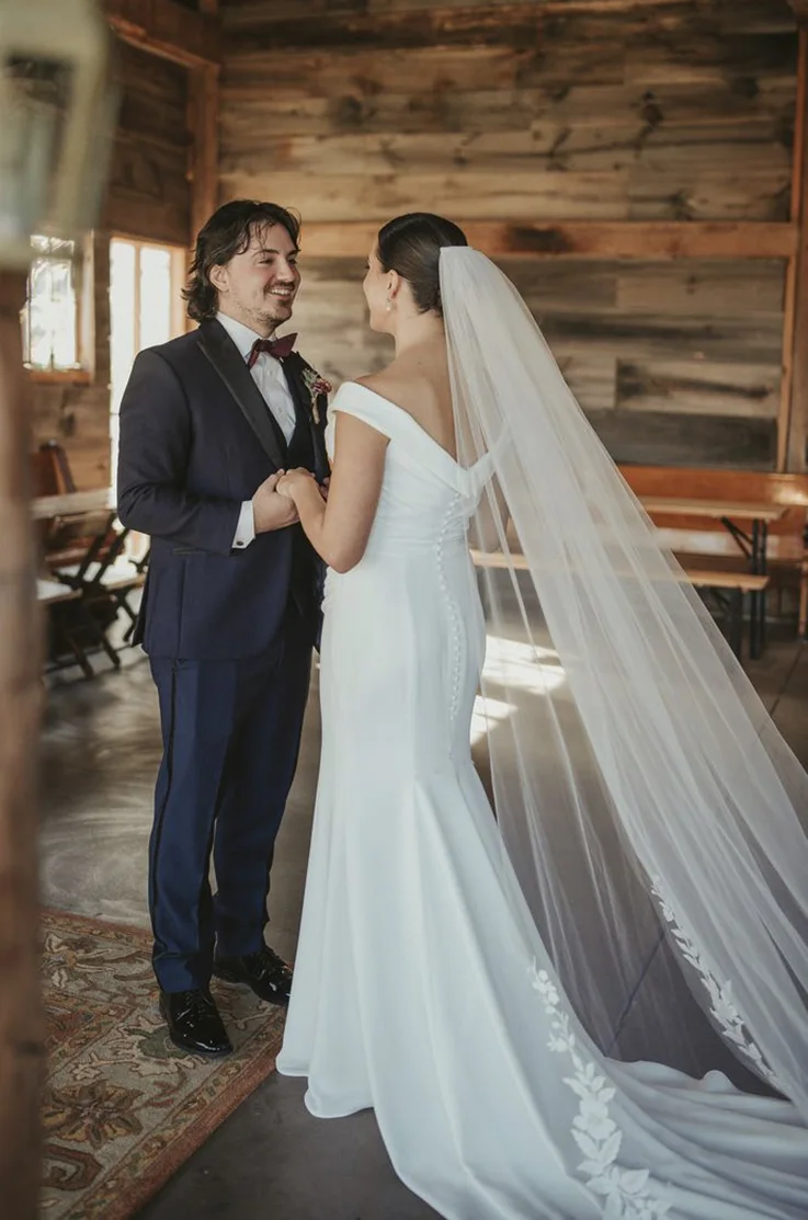 Bride in a crepe fit and flare wedding gown with a lace-trim veil, faces groom in a blue suit in a rustic barn setting.