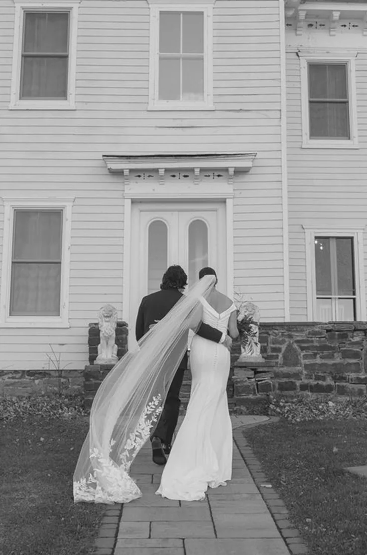 Bride in a fit and flare crepe wedding dress with an off-shoulder V-neckline, walks alongside her groom toward the venue.