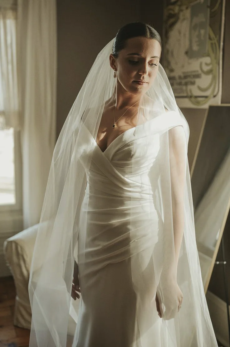 Close-up of bride in an off-shoulder draped gown with a crepe fit and flare skirt and a veil, posing indoors by a window. 