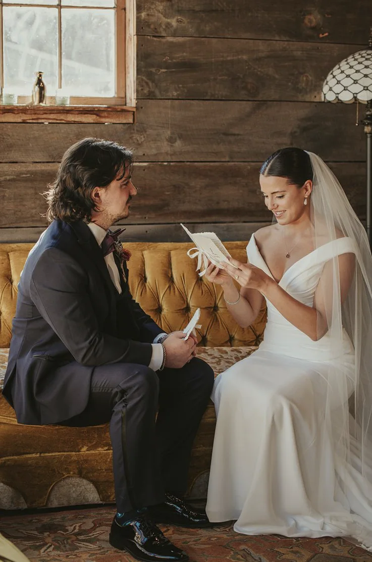 Bride in an off-shoulder draped crepe gown with fit and flare skirt and a veil, sits on a sofa reading vows with her groom.