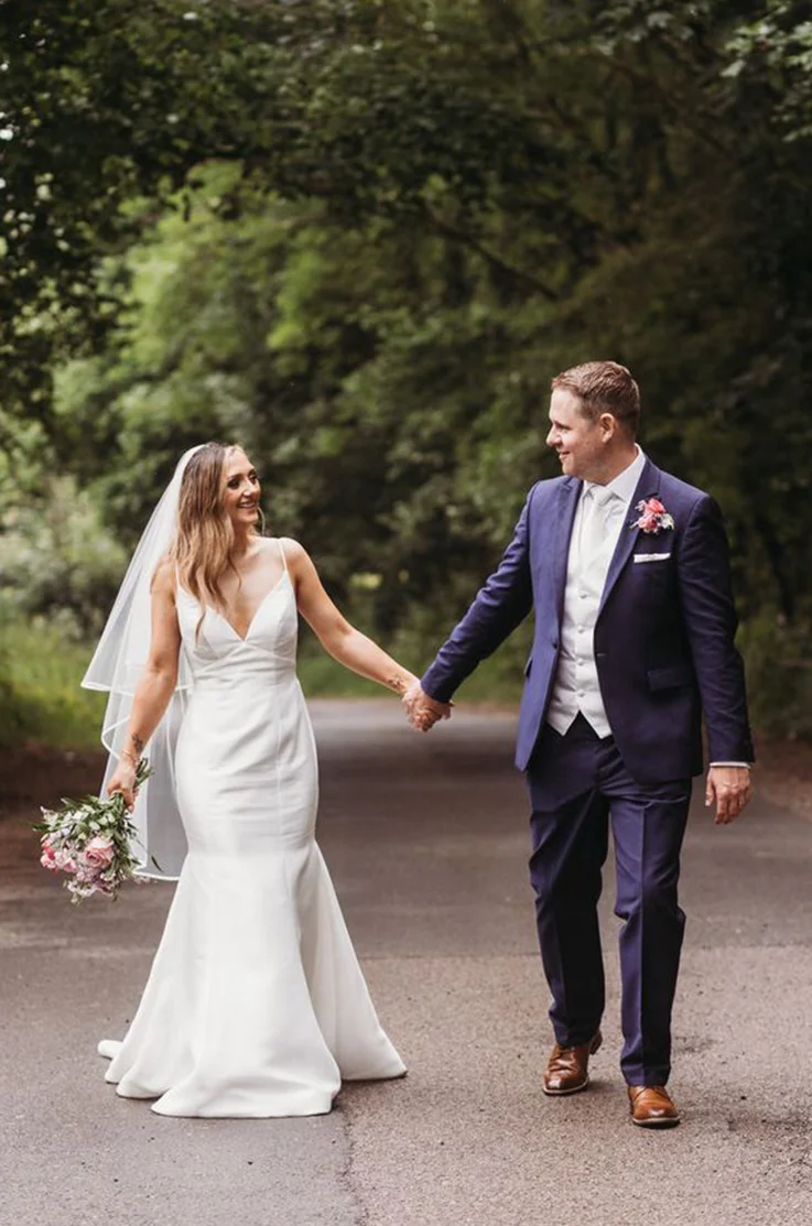 Bride in a fit and flare Mikado gown with a V-neckline and veil, holds hands with groom on an open road with trees.