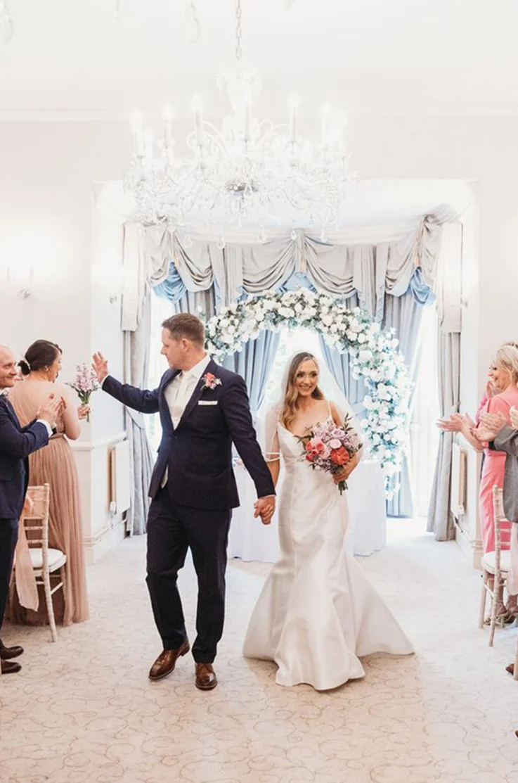 Bride in a princess seam Mikado wedding dress with a V-neck and veil, walks hand in hand with groom down the aisle.