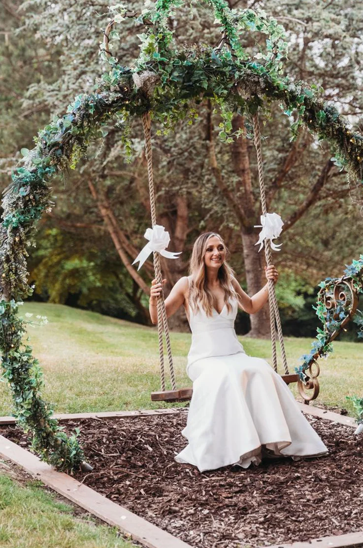 Bride in a V-neck fit and flare gown sits on a rope swing under a floral arch and trees outdoors, in an open field.