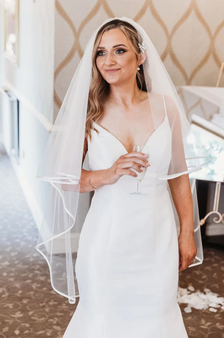 Close-up of bride in a Mikado fit and flare gown with a V-neckline and veil smiles indoors, holding champagne.
