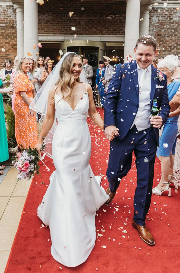 Bride in a V-neck Mikado gown with fit and flare skirt and a veil, holds a bouquet and hands with groom as they exit.