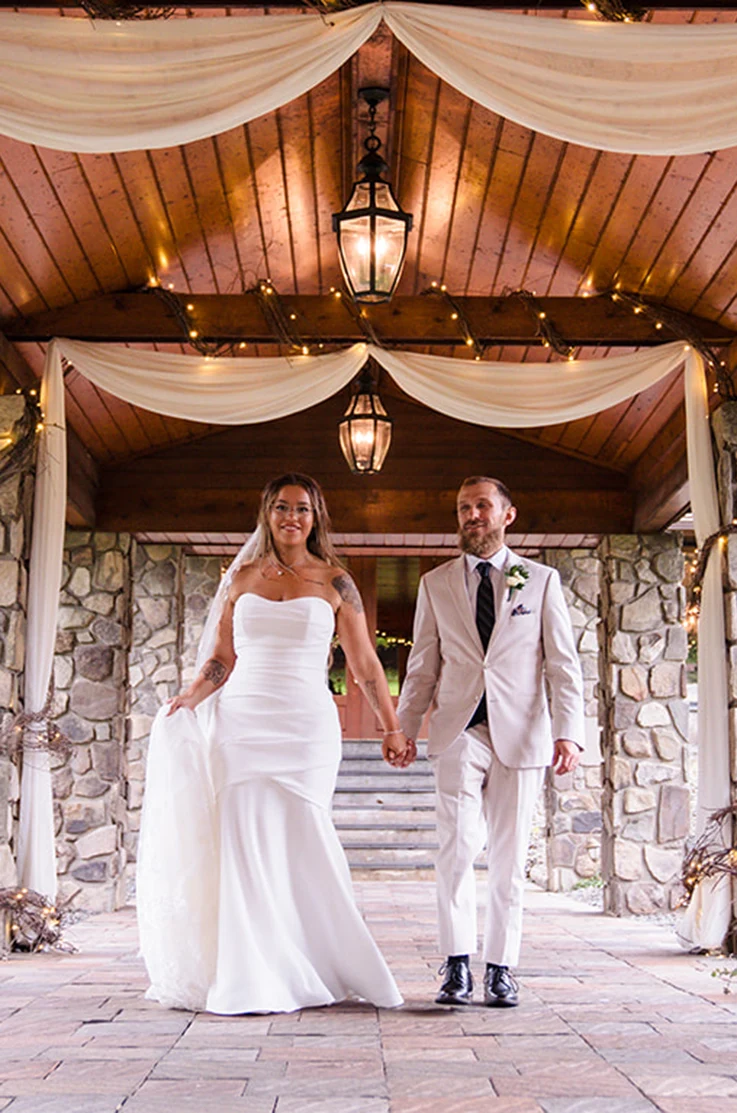Bride in a strapless crepe draped bodice wedding dress, holding hands with groom in a light gray suit under a pavilion