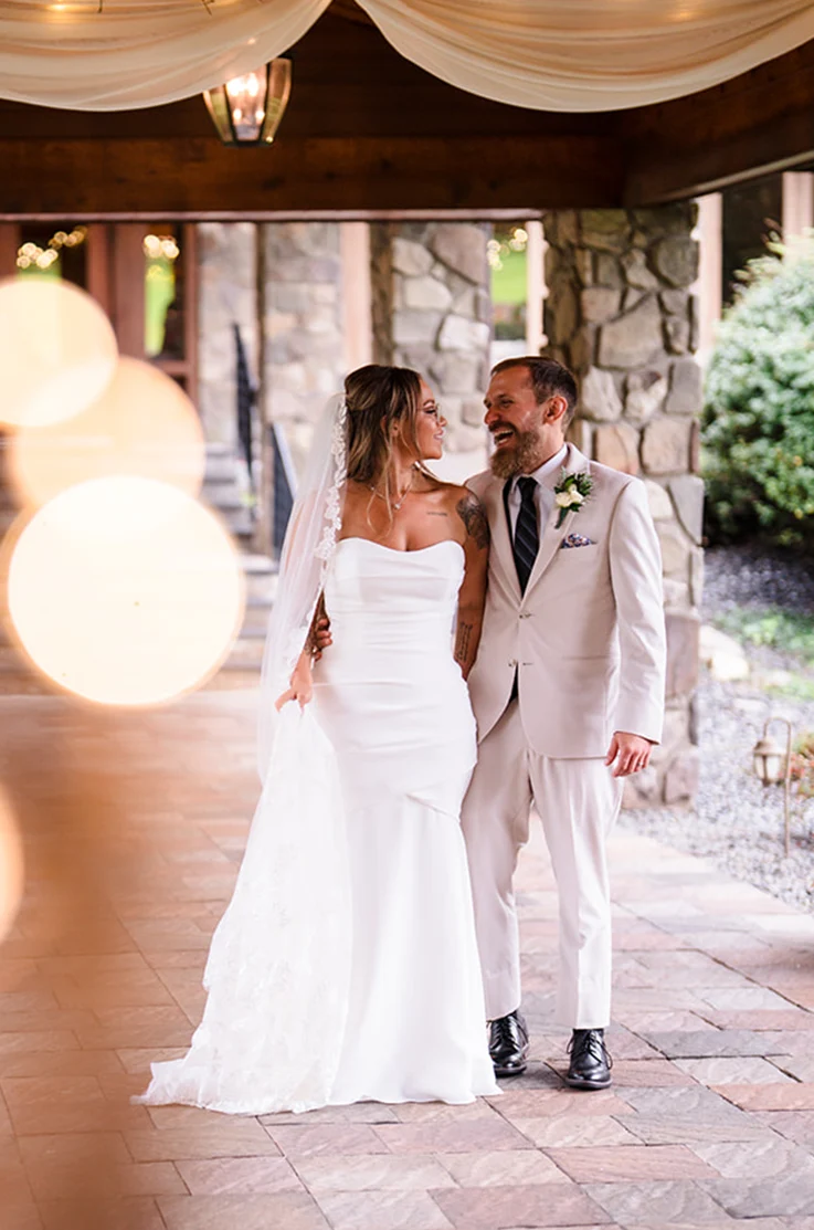 Groom embraces bride in a strapless ruched gown with a lace-trim veil, laughing on stone pavilion.