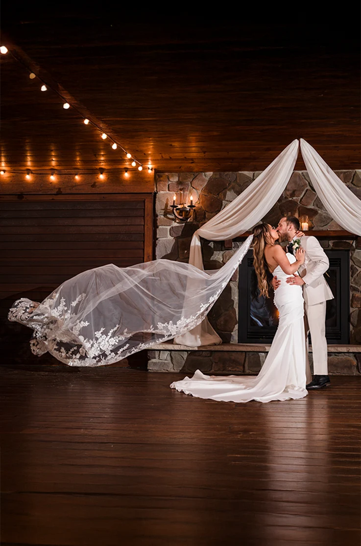Groom embraces bride in a strapless ruched crepe gown with a flowing veil, and they kiss near stone fireplace.