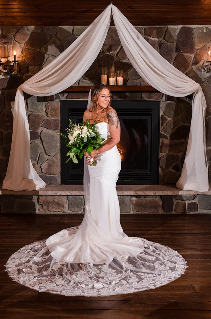 Bride poses in a crepe draped bodice gown with fit and flare skirt and a lace-trim veil, in front of a stone fireplace indoors.
