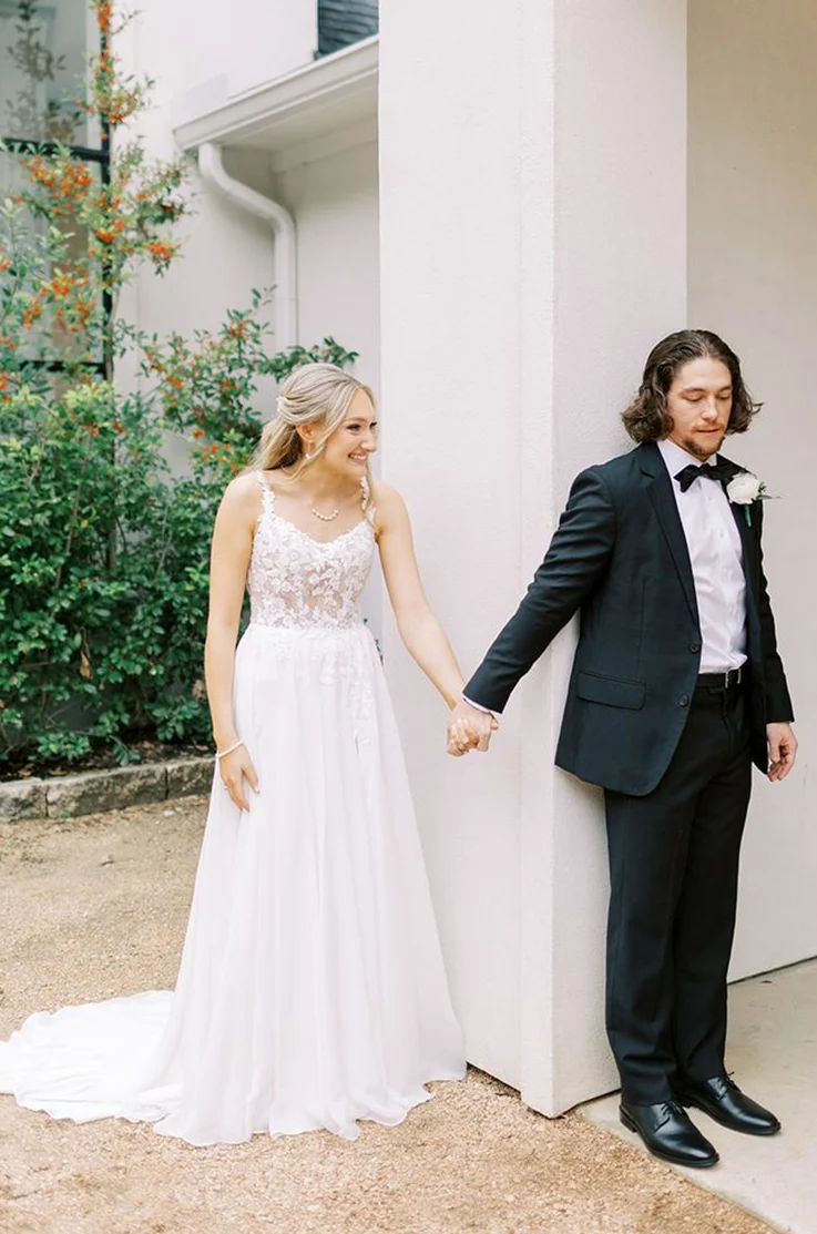 Bride in a scoop neck lace gown with a full organza skirt, smiles at groom as they hold hands outside the venue.