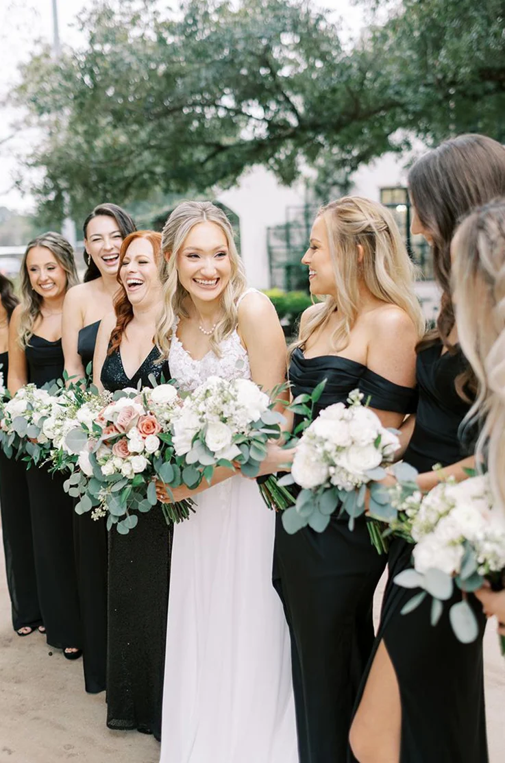 Bride smiles in a lace and organza gown with a scoop neckline, alongside bridesmaids in black dresses holding bouquets. 