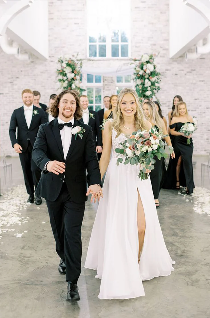 Bride in a lace bodice gown with an organza slit skirt, walks hand in hand with her groom down the aisle, holding a large bouquet.