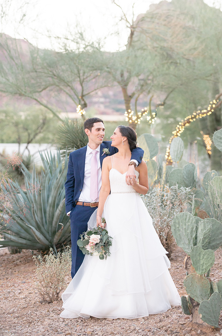 A groom in a suit has his arm around the shoulder of a bride in a ball gown wedding dress as they stand outside in a stone cactus garden, with string lights on trees behind them.