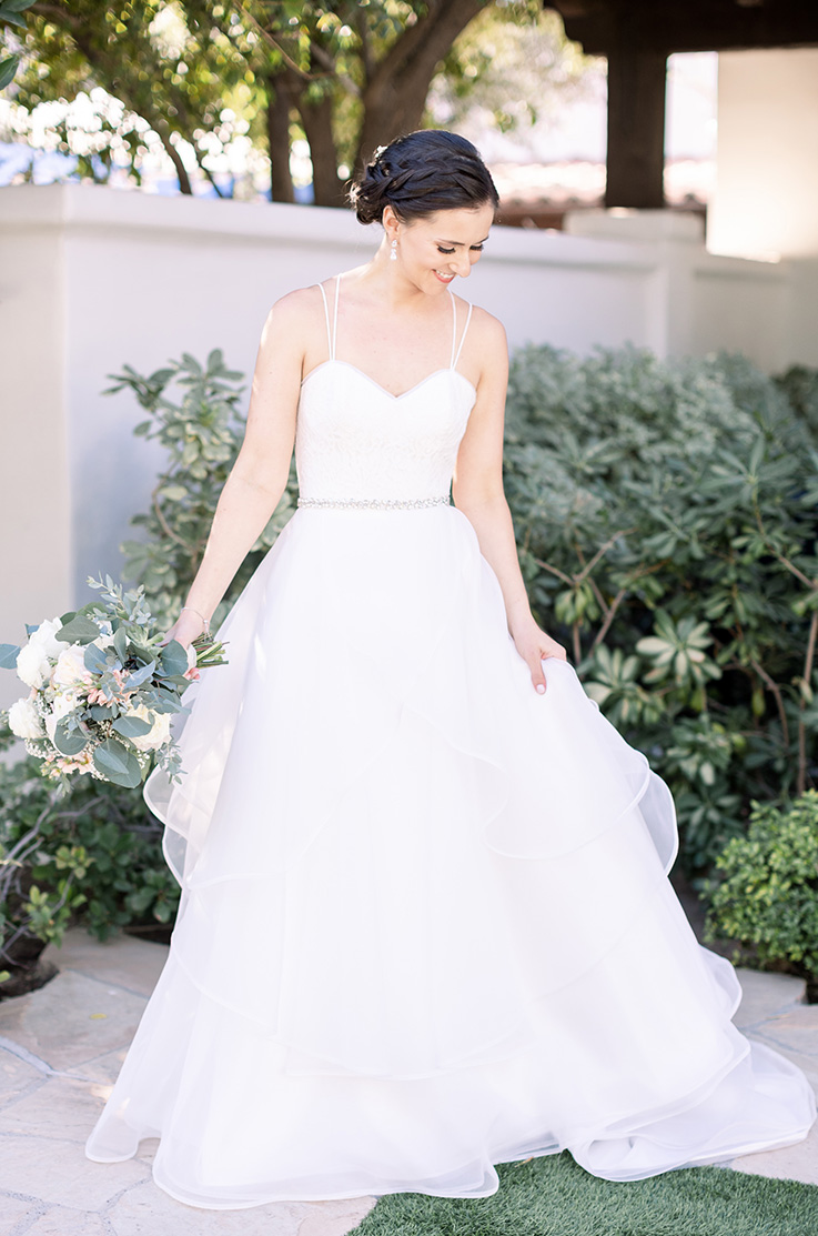 A bride in a ball gown wedding dress holds a bouquet and smiles while looking down, a she stands in a garden in front of greenery and concrete fencing.