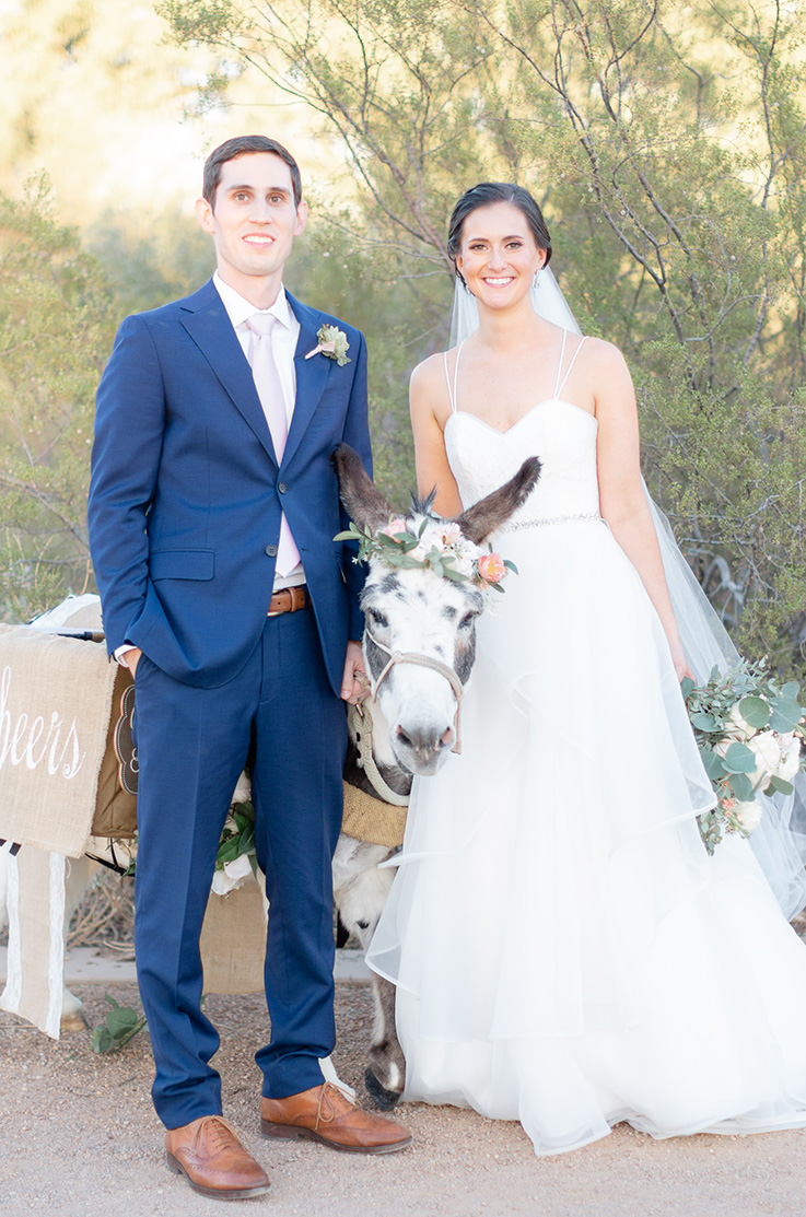 A bride in a ball gown wedding dress and a groom in a suit stand outside with a patient donkey wearing a flower crown, all of them standing on a gravel path with trees in the background.