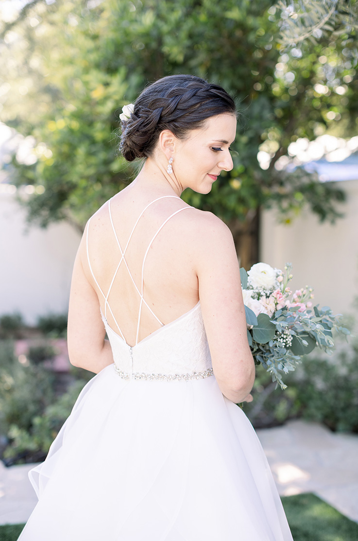 A bride in a ball gown wedding dress holds a bouquet and looks over her shoulder, while she stands outside in a garden, with a tree and bushes in the background.