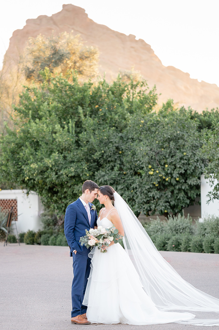 A bride in a ball gown wedding dress and a veil, holding a bouquet, and a groom in a suit, stand in a lot surrounded by bushes and trees, with a red rock mesa in the distance behind them.