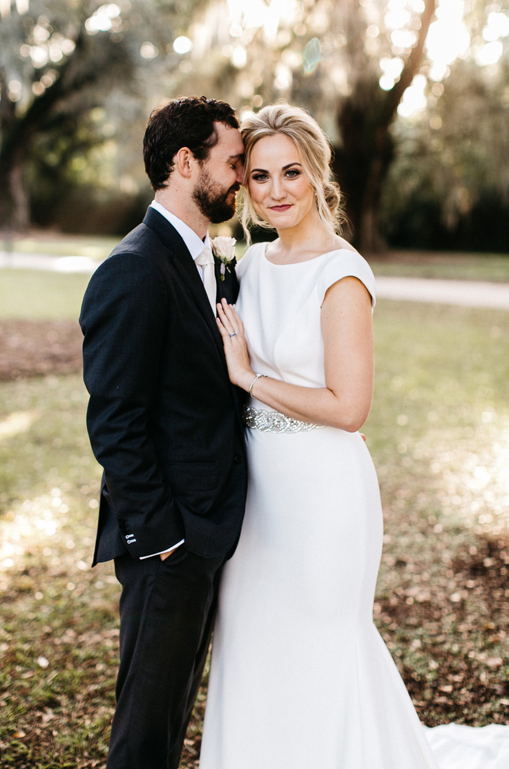 A bride in an embellished wedding dress and a groom in a suit smile and stand next to each other outside, with trees in the background.