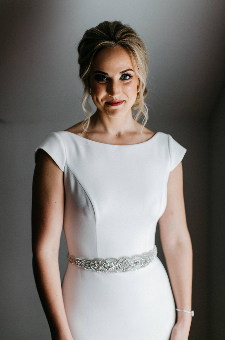A bride in an embellished cap sleeve wedding dress stands indoors in front of a blank wall and smiles.