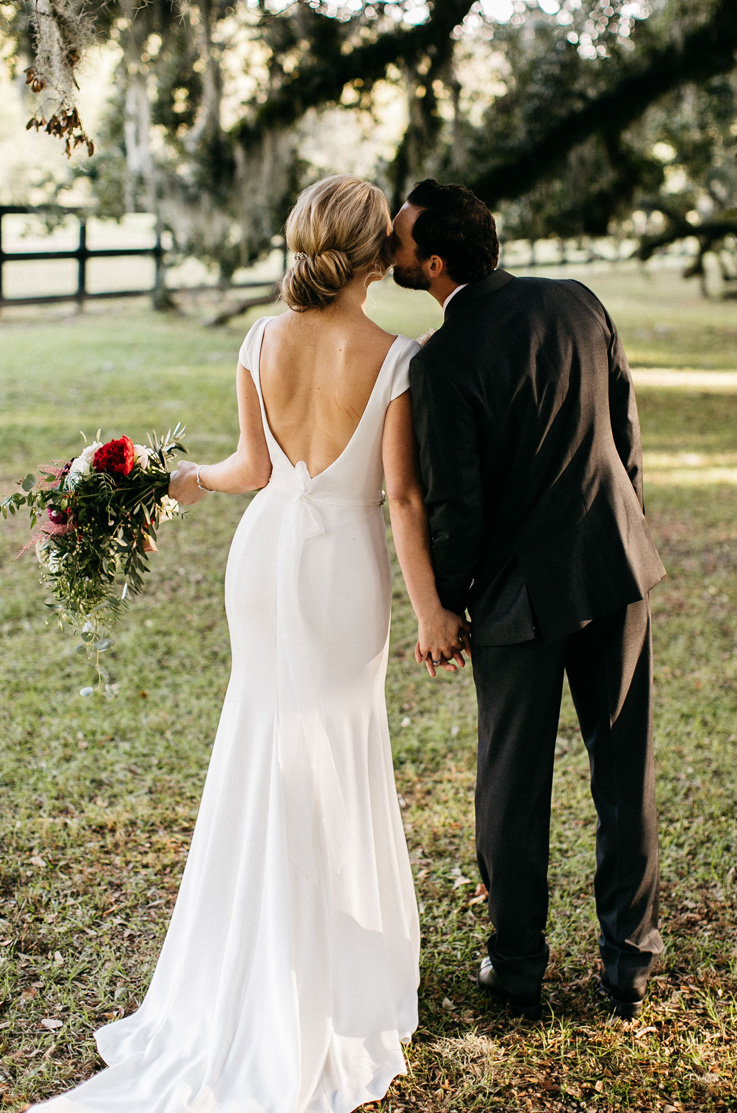 A groom in a suit kisses the cheek of a bride in a cap sleeve wedding dress while they hold hands, the bride has a bouquet in her other hand. They are standing outside in the grass, with tree branches above them.