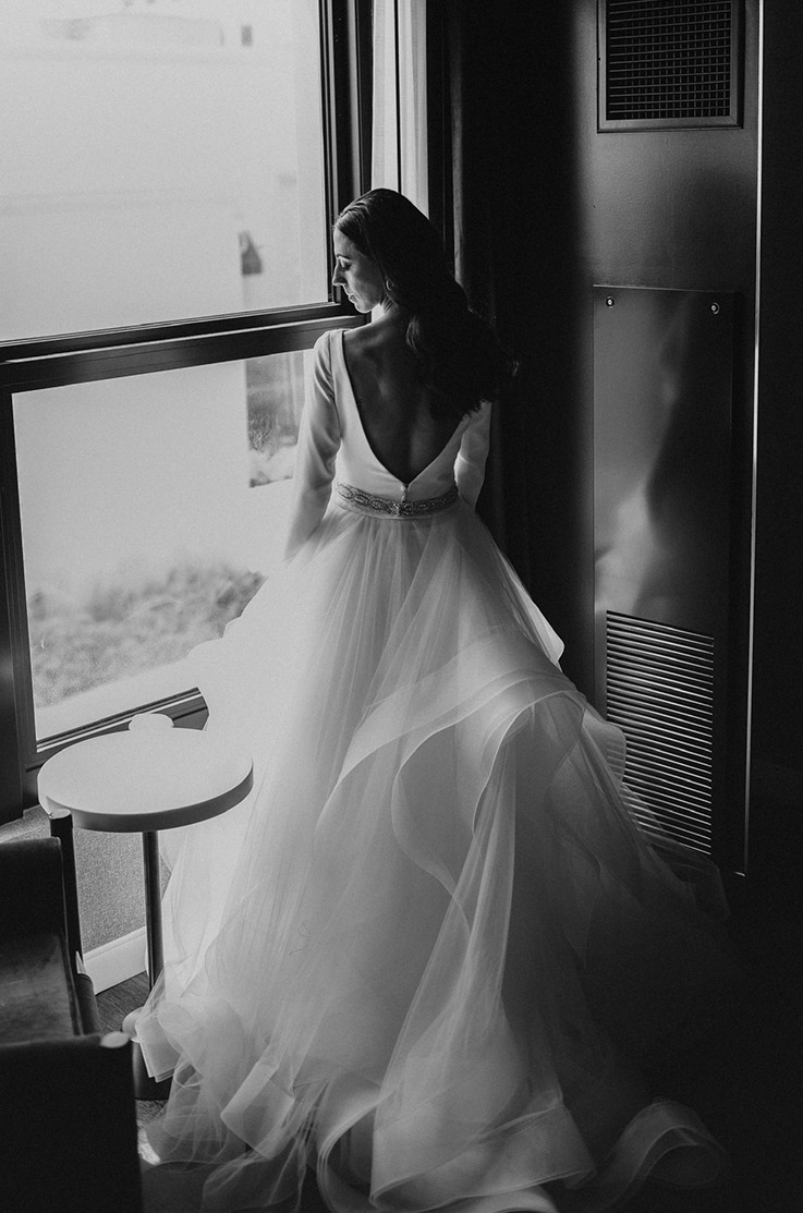 A bride in a long sleeve wedding dress stands indoors in front of a large window, while looking down over her shoulder.