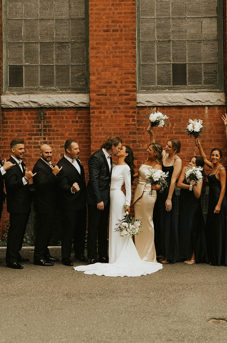 A bride in a long sleeve wedding dress and a groom in a suit kiss outside of a brick building while their wedding party cheers for them.