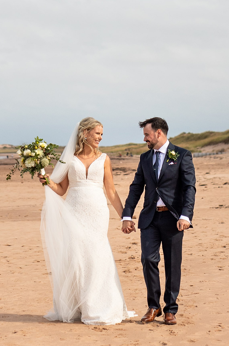 A bride in a sleeveless lace wedding dress and a veil holds a bouquet while holding hands with a groom in a suit, both walking along a sandy beach, looking at each other and smiling.