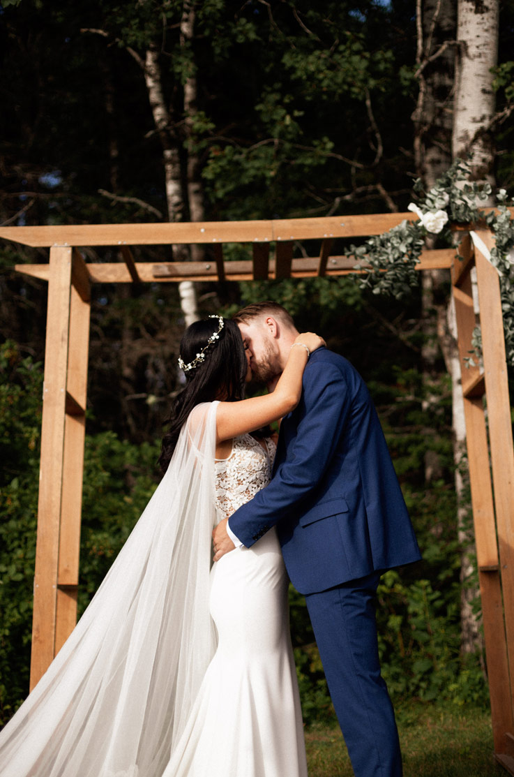 A bride in a wedding dress and a tulle cape, and a groom in a suit, kiss outside under a decorated wooden arbor.