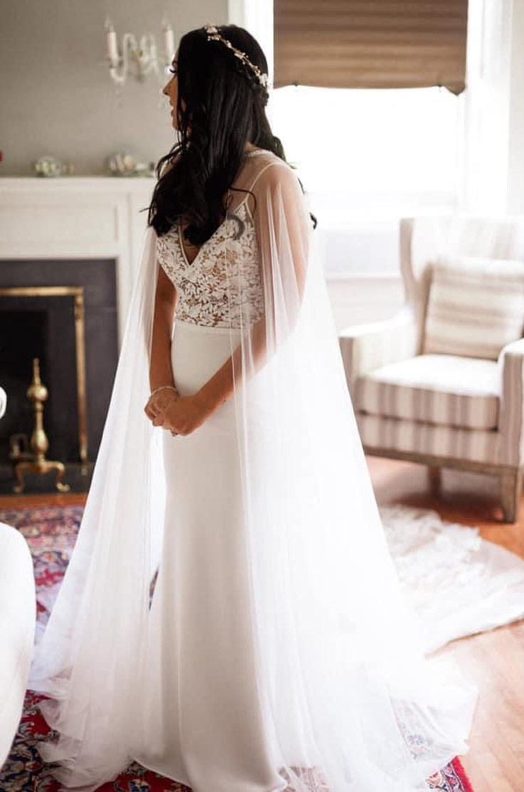 A bride in a V-neck wedding dress and a tulle cape stands indoors and looks away over her shoulder, with a chair and fireplace in the background.