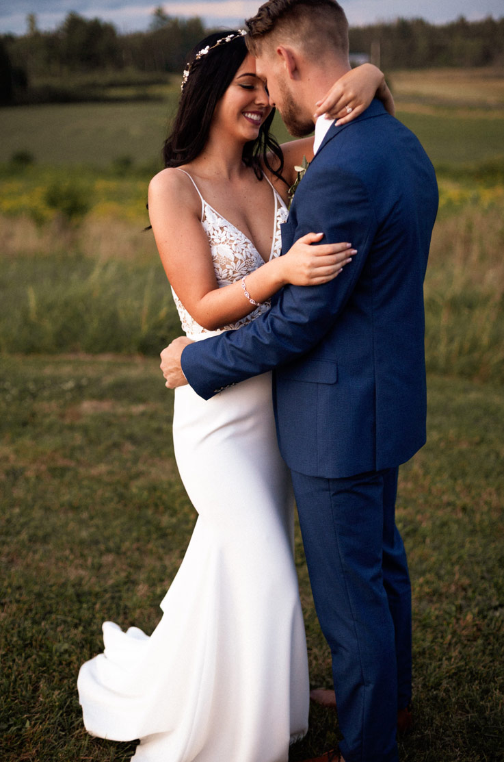 A smiling bride in a V-neck wedding dress and a groom in a suit hold each other while standing outside in a grassy field.