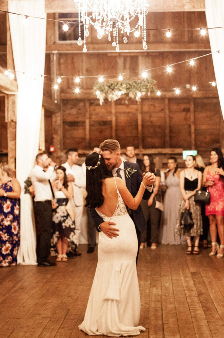 A bride in a bustled wedding dress and a groom in a suit dance in a decorated barn with a chandelier overhead, while wedding guests watch in the background.