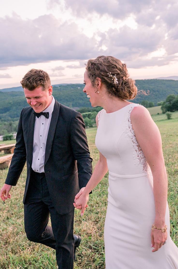 A bride in a sleeveless wedding dress and a groom in a suit walk hand in hand in a grass field while smiling and laughing.