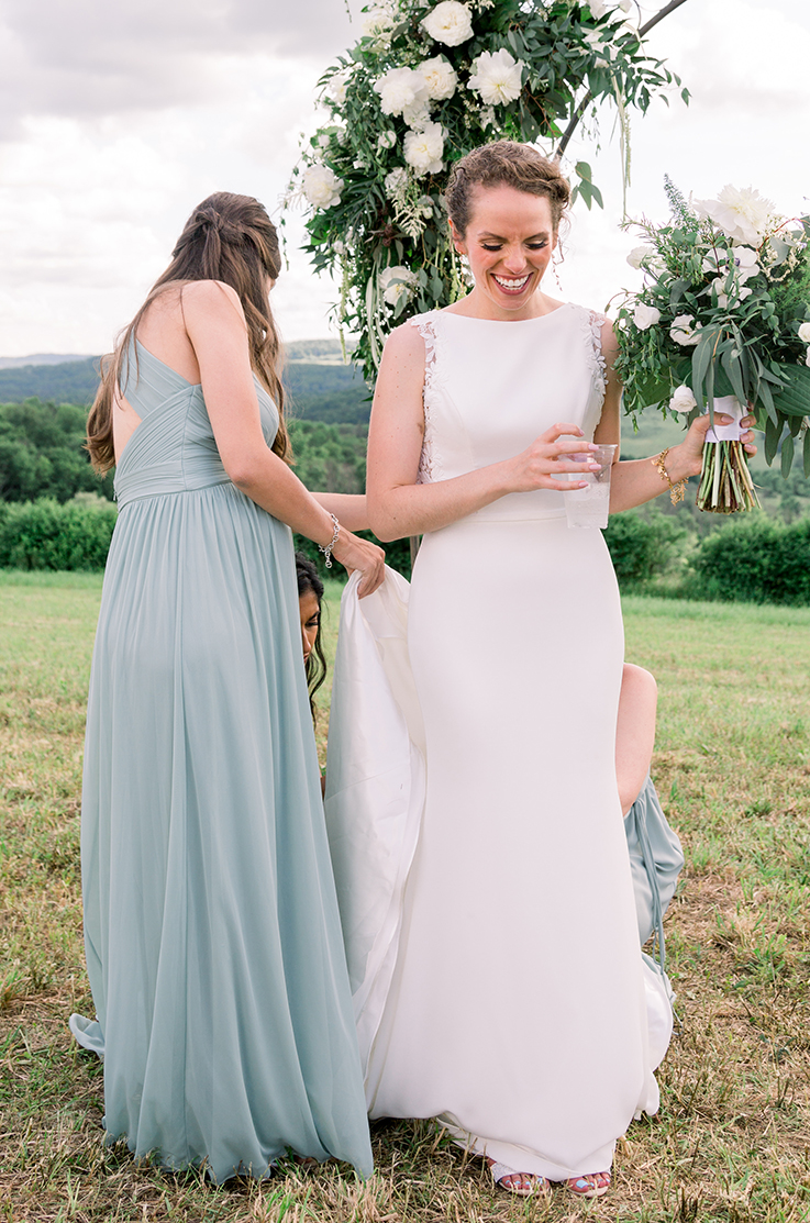 A smiling bride in a sleeveless wedding dress holds a bouquet in one hand and a cup in the other, while bridesmaids fix her train in front of a wedding ceremony decoration, all standing in a field of grass outdoors.