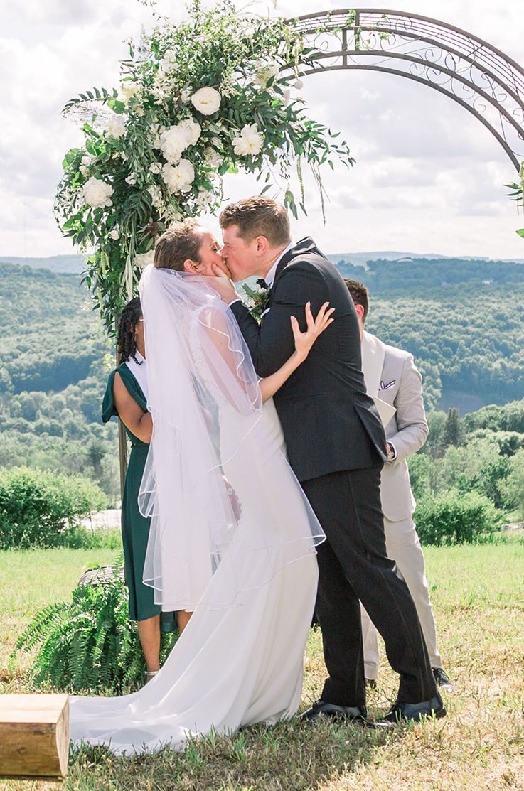 A groom in a suit kisses a bride in a sleeveless wedding dress and veil in front of a decorated wedding arc in a grass field outdoors, with rolling hills in the background.