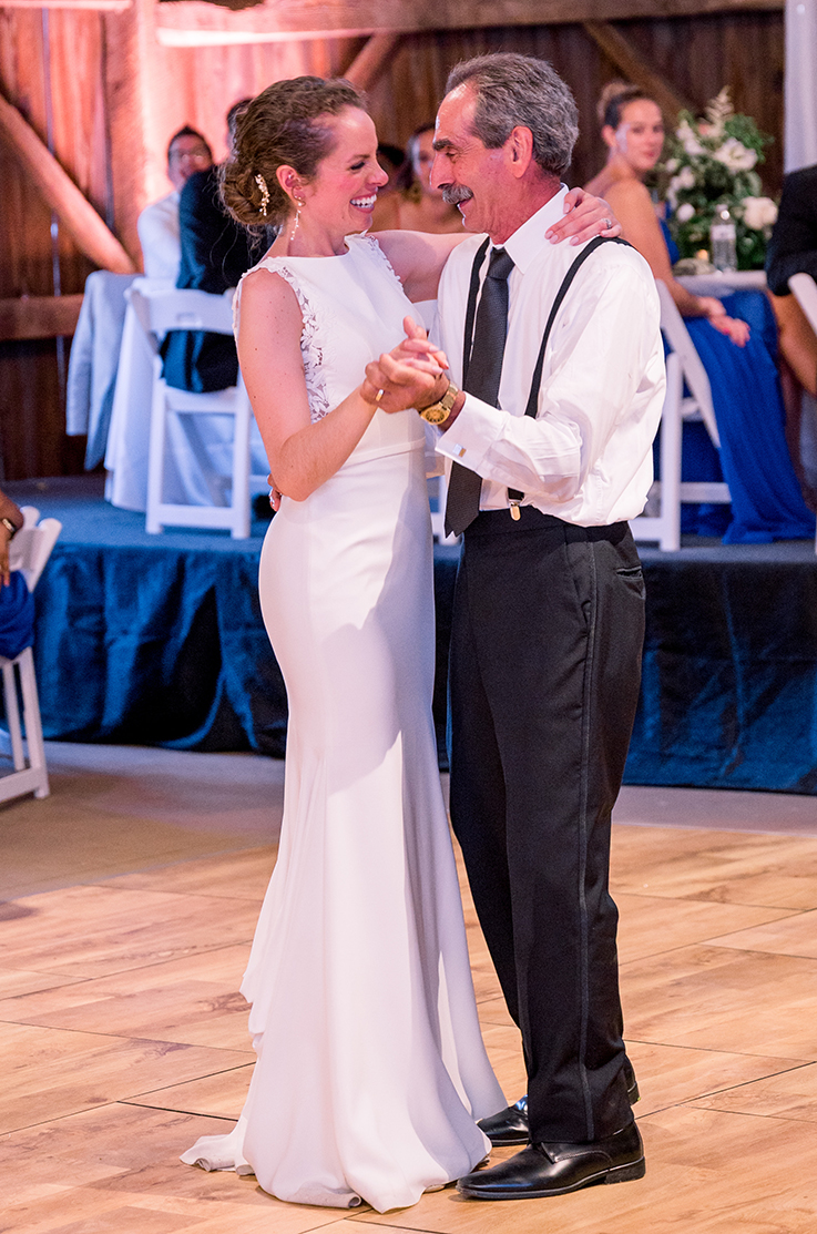 A bride in a sleeveless wedding dress and a man in dress pants and suspenders dance indoors on a dance floor, while wedding guests sitting in chairs watch.