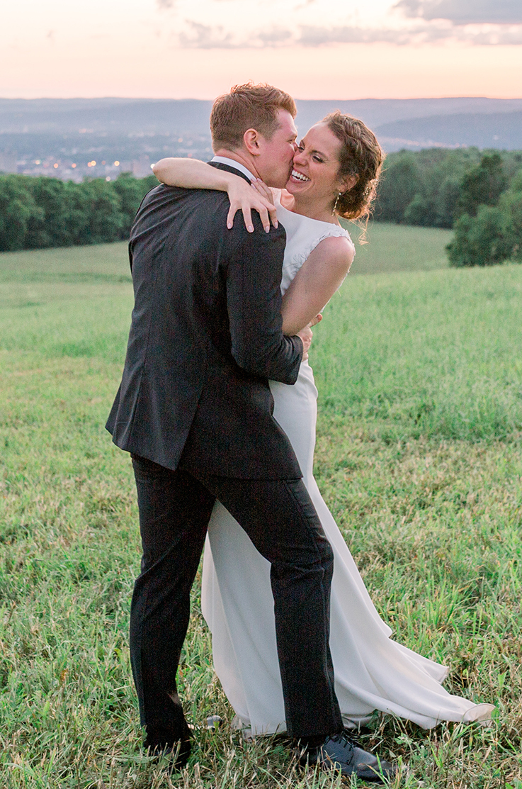 A groom in a suit kisses a bride in a sleeveless wedding dress while she laughs and holds the groom, both of them standing outside in a grass field.