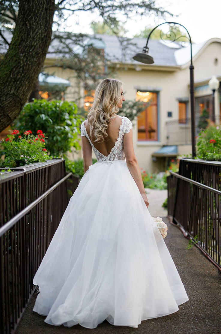 A bride in a cap sleeve wedding dress stands facing away on a pedestrian bridge, in front of a house and garden.