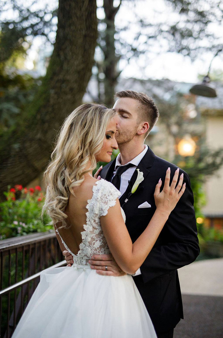 A groom in a suit kisses the forehead of a bride in a cap sleeve wedding dress while they stand outdoors, with a tree and building in the background.