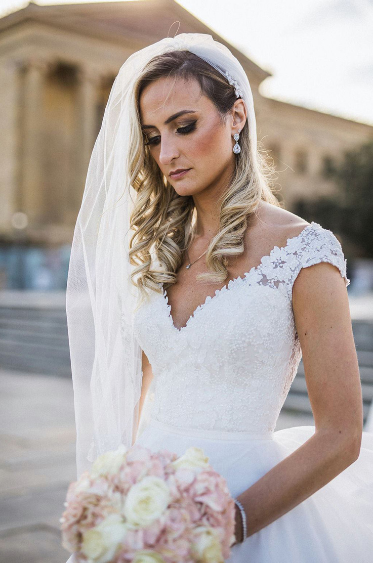 A bride in a cap sleeve wedding dress and veil holds a bouquet and looks down, while standing outside in front of a pillared building.