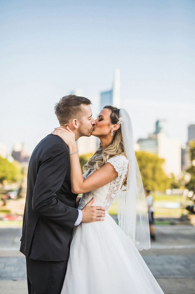 A bride in a cap sleeve wedding dress and veil, and a groom in a suit kiss while standing outside, with a city in the distance in the background.
