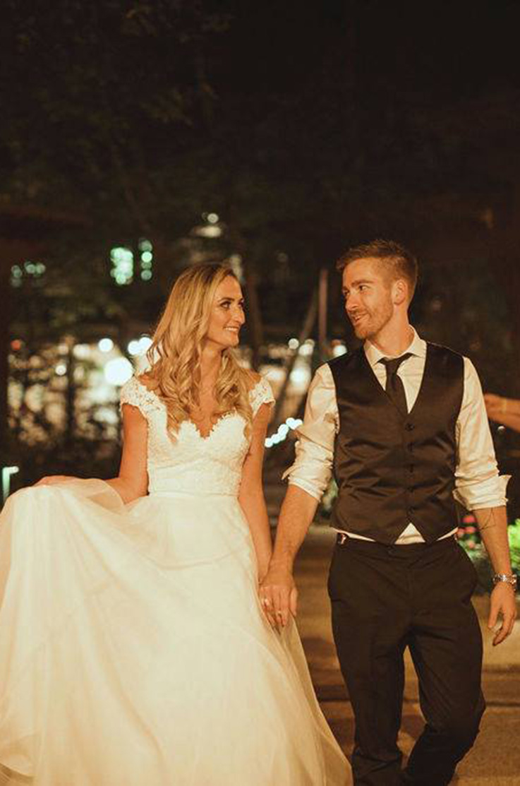 A bride in a cap sleeve wedding dress and a groom in a suit vest walk together outside at night while looking at each other and smiling.